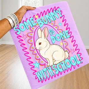 Może przedstawiać: Jasnofioletowy koc z napisem "SOME BUNNY IS DONE WITH SCHOOL!" i ilustracją białego kr&oacute;lika. Projekt zawiera r&oacute;żowe i złote akcenty z gwiazdami.