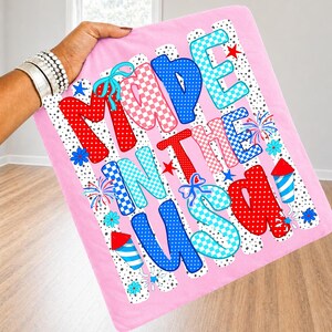 Pode incluir: Um item de tecido rosa de formato quadrado com o texto MADE IN THE USA em letras grandes, multicoloridas e estampadas. O design inclui bolinhas vermelhas, azuis e brancas, padr&otilde;es xadrez, ilustra&ccedil;&otilde;es de fogos de artif&iacute;cio e flores azuis.
