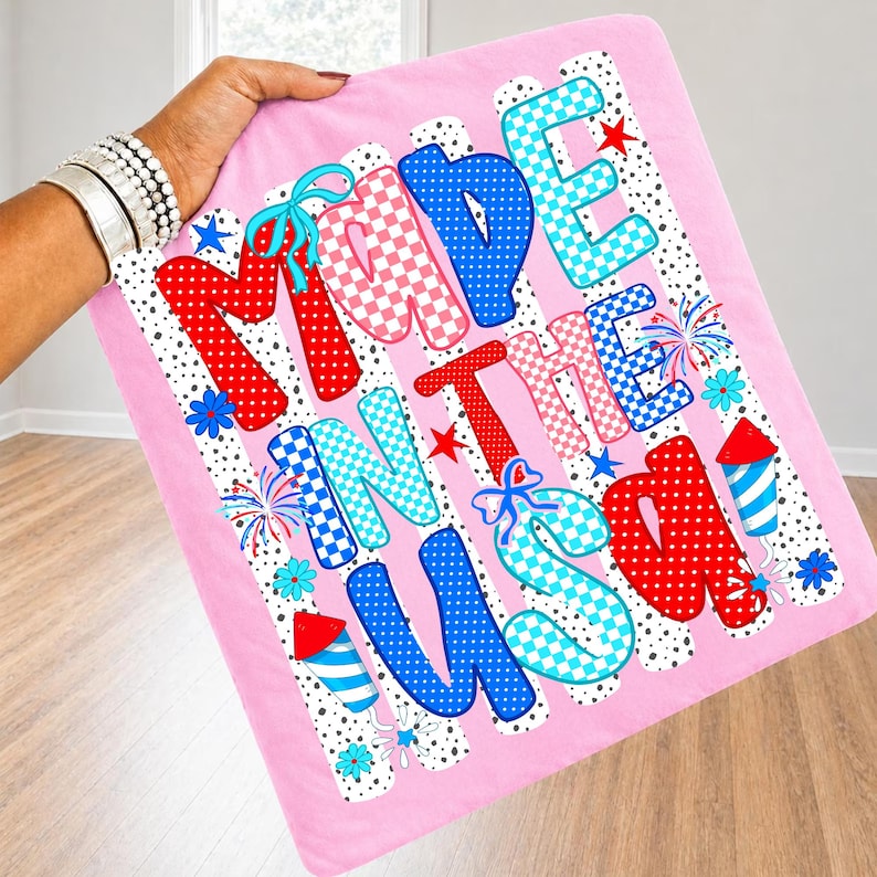 Pode incluir: Um item de tecido rosa de formato quadrado com o texto MADE IN THE USA em letras grandes, multicoloridas e estampadas. O design inclui bolinhas vermelhas, azuis e brancas, padr&otilde;es xadrez, ilustra&ccedil;&otilde;es de fogos de artif&iacute;cio e flores azuis.