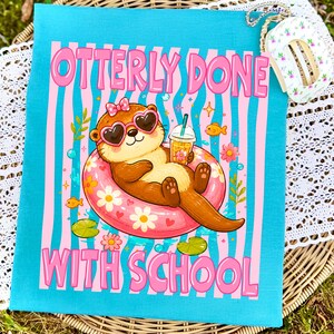 K&ouml;nnte beinhalten: Ein t&uuml;rkisfarbenes Handtuch mit einem Cartoon-Otter, der eine herzf&ouml;rmige Sonnenbrille tr&auml;gt und in einem rosa Schwimmring schwimmt. Der Text "OTTERLY DONE WITH SCHOOL" ist rosa. Der Otter h&auml;lt ein Getr&auml;nk mit einem Strohhalm.