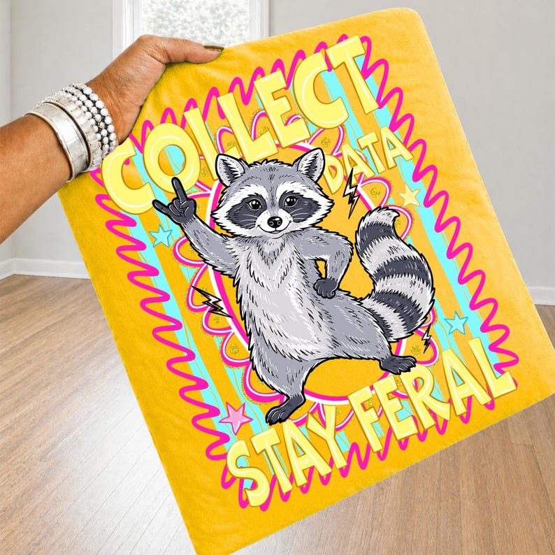 May include: Yellow blanket featuring a cartoon raccoon illustration. The raccoon is grey and white, giving the middle finger. Text on the blanket reads "COLLECT DATA STAY FERAL" in yellow and pink. The blanket is held by a person.