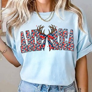 Pu&ograve; includere: T-shirt azzurra con la scritta "AMERICA" in un motivo mimetico rosso, bianco e blu. Un fiocco e delle corna sono centrati sopra la parola. La maglietta &egrave; uno stile casual.