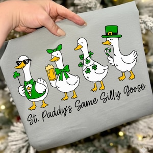May include: A light grey graphic featuring four cartoon geese celebrating St. Patrick's Day. The geese are adorned with shamrocks, bow ties, and a green hat. The text "St. Paddy's Same Silly Goose" is written below the geese in a playful font.