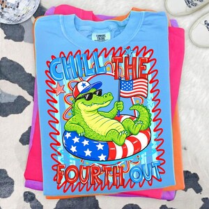 K&ouml;nnte beinhalten: Hellblaues T-Shirt mit einem Aufdruck eines Alligators mit Sonnenbrille und patriotischem Hut, der auf einem rot-wei&szlig;-blauen Schwimmring schwimmt. Der Text lautet "Chill the Fourth Out."