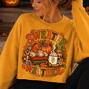 Puede incluir: Una sudadera de manga larga color amarillo mostaza con un estampado gr&aacute;fico de una calabaza, una pila de mantas dobladas, un termo y una taza con vapor. El texto dice "SWEATER WEATHER" y "GOOD THINGS AHEAD" con hojas de oto&ntilde;o y bellotas.