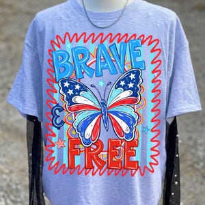 May include: A light grey t-shirt with a patriotic butterfly design in red, white, and blue. The words "BRAVE" and "FREE" are in blue and red, respectively, with a wavy border. The butterfly has stars on its wings.