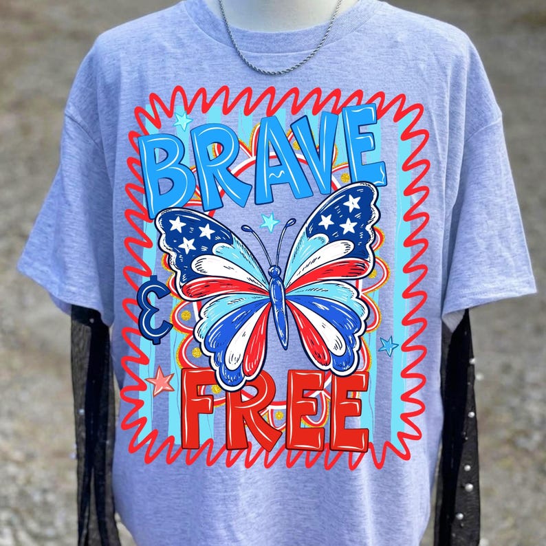 May include: A light grey t-shirt with a patriotic butterfly design in red, white, and blue. The words "BRAVE" and "FREE" are in blue and red, respectively, with a wavy border. The butterfly has stars on its wings.