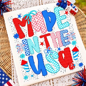 Pode incluir: Uma t-shirt branca com o texto MADE IN THE USA em letras grandes, coloridas e estampadas. O design inclui padr&otilde;es de bolinhas e xadrez em vermelho, azul e azul claro, acentuados com pequenas flores azuis, estrelas vermelhas e fogos.