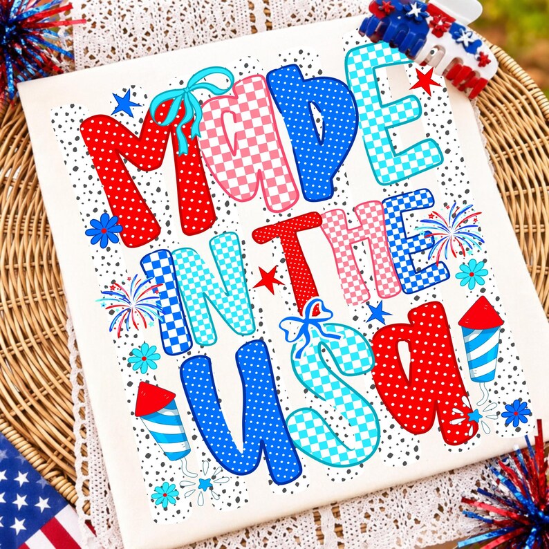 Pode incluir: Uma t-shirt branca com o texto MADE IN THE USA em letras grandes, coloridas e estampadas. O design inclui padr&otilde;es de bolinhas e xadrez em vermelho, azul e azul claro, acentuados com pequenas flores azuis, estrelas vermelhas e fogos.