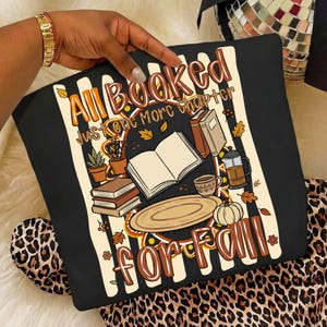 K&ouml;nnte beinhalten: Ein schwarzes Sweatshirt mit einem Grafikdruck und dem Text All Booked Just one More chapter FOR FALL. Das Design zeigt Illustrationen von offenen und gestapelten B&uuml;chern, einer Kaffeepresse, einer Tasse, einem K&uuml;rbis und Topfpflanzen auf Streifen.