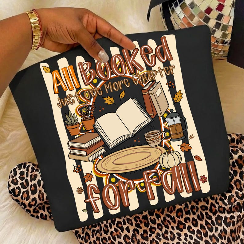 K&ouml;nnte beinhalten: Ein schwarzes Sweatshirt mit einem Grafikdruck und dem Text All Booked Just one More chapter FOR FALL. Das Design zeigt Illustrationen von offenen und gestapelten B&uuml;chern, einer Kaffeepresse, einer Tasse, einem K&uuml;rbis und Topfpflanzen auf Streifen.