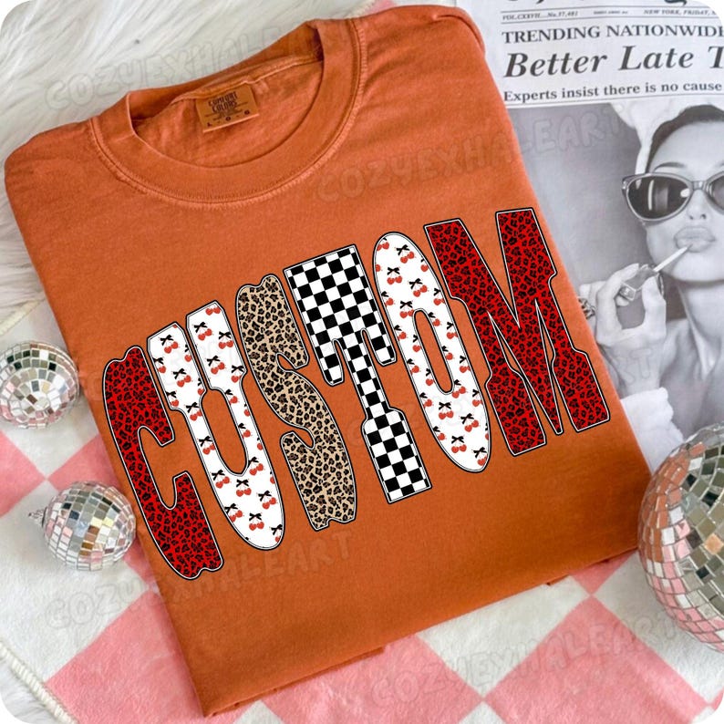 May include: An orange t-shirt with the word "CUSTOM" in a bold, stylized font. The letters feature various patterns, including red leopard print, cherries, and a black and white checkerboard design. The shirt is folded and laid flat.