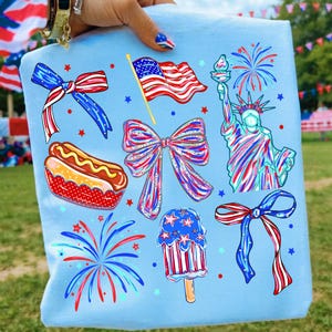 May include: A light blue tote bag with patriotic designs. The illustrations include a waving American flag, fireworks, the Statue of Liberty, bows, a hot dog, and a popsicle, in red, white, and blue.