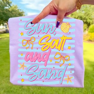 Peut inclure: Un t-shirt lilas avec l'inscription "Sun, Salt and Sand" dans une police de caract&egrave;res color&eacute;e et ludique. Le motif comprend des rayures bleues, des n&oelig;uds jaunes, des &eacute;toiles de mer et un coquillage. Le t-shirt est tenu par une personne.