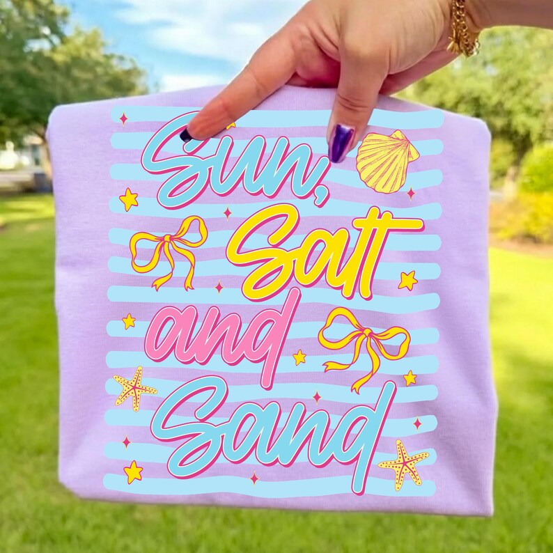Peut inclure: Un t-shirt lilas avec l'inscription "Sun, Salt and Sand" dans une police de caract&egrave;res color&eacute;e et ludique. Le motif comprend des rayures bleues, des n&oelig;uds jaunes, des &eacute;toiles de mer et un coquillage. Le t-shirt est tenu par une personne.