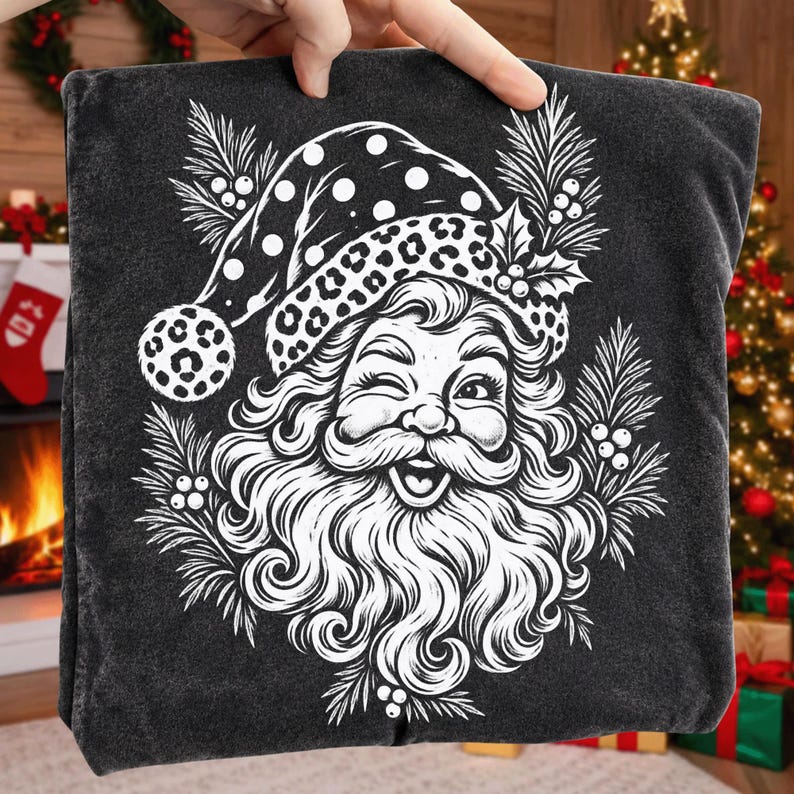 K&ouml;nnte beinhalten: Ein anthrazitfarbenes T-Shirt mit einem wei&szlig;en Grafikdruck eines zwinkernden Weihnachtsmanns, der eine M&uuml;tze mit Leopardenmuster tr&auml;gt. Das Design ist von Tannenzweigen und Stechpalmenbeeren umgeben und bildet eine festliche Illustration.