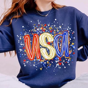Peut inclure: Sweat-shirt bleu marine avec le mot "USA" en lettres rouges, blanches et bleues. Les lettres sont bord&eacute;es d'or et entour&eacute;es d'un motif &eacute;clabouss&eacute; de rouge, blanc, bleu et or. Un sweat-shirt de style d&eacute;contract&eacute;.