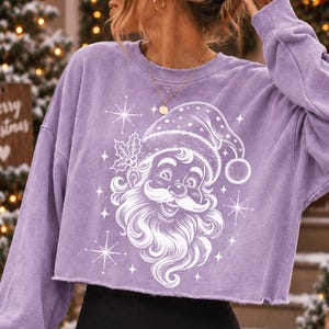 May include: A light purple long-sleeve cropped sweatshirt featuring a white graphic print of a smiling Santa Claus wearing a hat with holly leaves. The design includes several four-pointed star accents surrounding the central Santa Claus illustration.