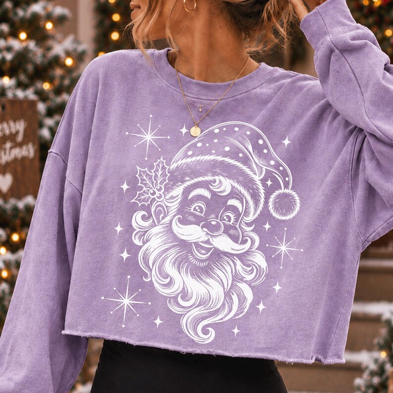 May include: A light purple long-sleeve cropped sweatshirt featuring a white graphic print of a smiling Santa Claus wearing a hat with holly leaves. The design includes several four-pointed star accents surrounding the central Santa Claus illustration.
