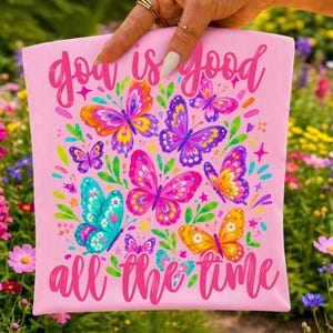 May include: A light pink fabric square featuring the text "god is good all the time" in a dark pink script font. The design includes multiple colorful butterflies in shades of orange, purple, pink, yellow, and teal, accented with green leaves.