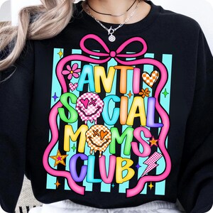 K&ouml;nnte beinhalten: Schwarzes Sweatshirt mit dem Text "ANTI SOCIAL MOMS CLUB" in einem farbenfrohen Design. Die Grafik enth&auml;lt ein rosa Band, Smiley-Gesichter, Blumen und einen Blitz, vor einem blau gestreiften Hintergrund.