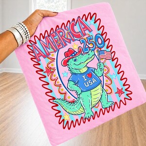 Puede incluir: Manta rosa con un cocodrilo de dibujos animados con sombrero de vaquero y camiseta azul con "I ❤️ USA". El cocodrilo sostiene una peque&ntilde;a bandera americana. La manta tiene el texto "AMERICA 250" en rojo y azul.