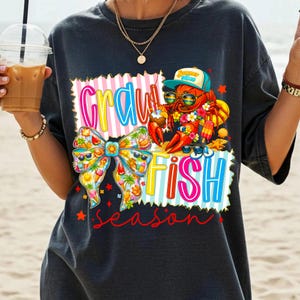 May include: Dark grey t-shirt with a colourful graphic of a lobster wearing sunglasses and a hat, holding a coconut drink. The words "Crawfish Fish Season" are displayed in a vibrant font, with a bow and other summer-themed elements.