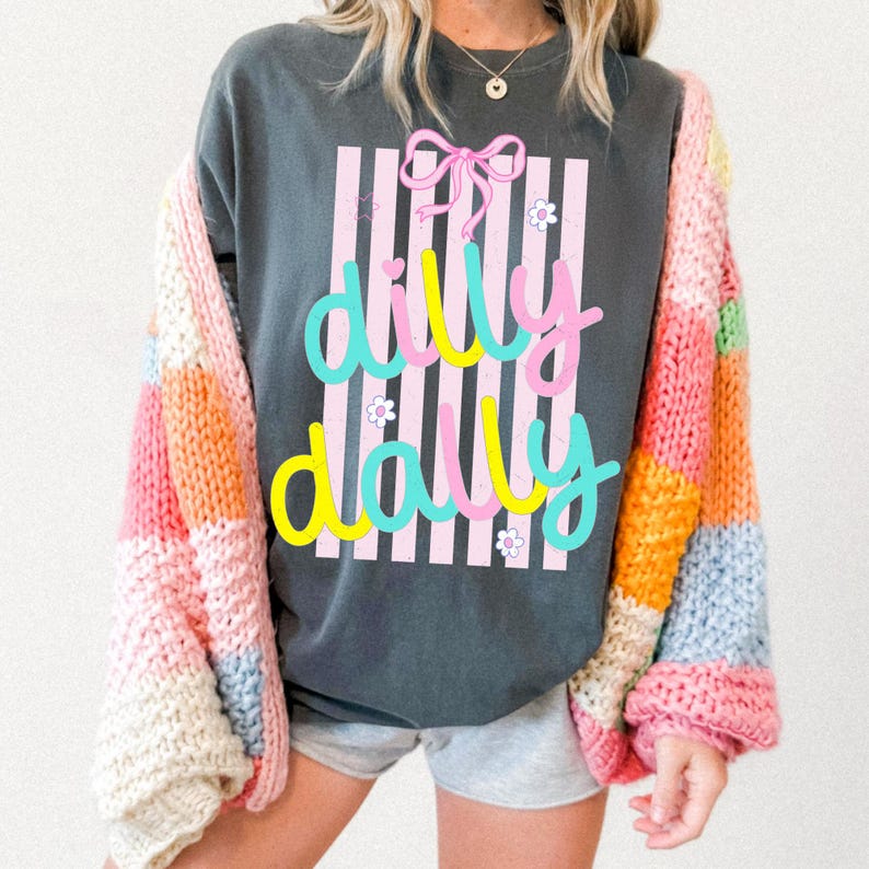 May include: Dark grey oversized t-shirt featuring the words "dilly dally" in a colourful script, set against a pink and white striped background. A pink bow and small white flowers accent the design. A multi-coloured, chunky knit cardigan is worn.
