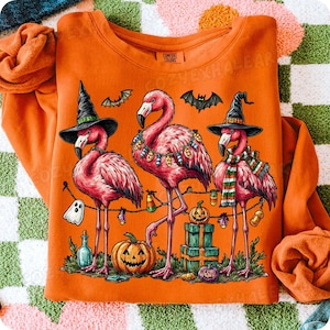 May include: Orange sweatshirt with a Halloween design. It features three pink flamingos wearing witch hats and scarves, decorated with lights and pumpkins. Bats and a jack-o'-lantern are also included in the illustration.