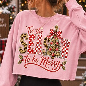 Op de afbeelding: Een lichtroze cropped sweatshirt met lange mouwen en een grafische print met de tekst 'Tis the SEASON to be Merry'. Het ontwerp bevat een kerstboom met luipaardprint, een rode strik met witte stippen, rood-wit geblokte letters en hulst.