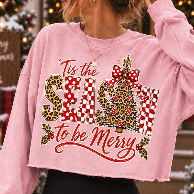 Op de afbeelding: Een lichtroze cropped sweatshirt met lange mouwen en een grafische print met de tekst 'Tis the SEASON to be Merry'. Het ontwerp bevat een kerstboom met luipaardprint, een rode strik met witte stippen, rood-wit geblokte letters en hulst.