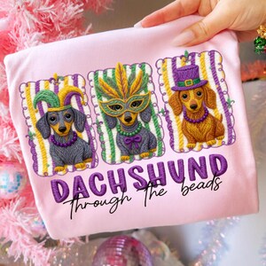 Puede incluir: Camiseta rosa claro con tres perros salchicha bordados con disfraces de Mardi Gras. Cada perro lleva un accesorio diferente: un gorro de buf&oacute;n, una m&aacute;scara de plumas y un sombrero de copa. El texto "DACHSHUND Through the beads" est&aacute; bordado debajo.
