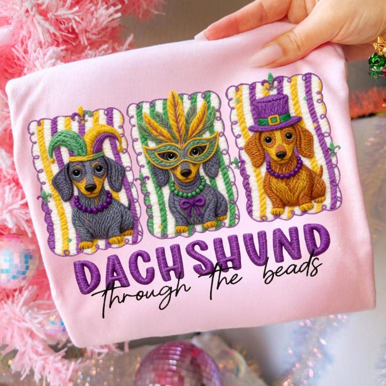 Puede incluir: Camiseta rosa claro con tres perros salchicha bordados con disfraces de Mardi Gras. Cada perro lleva un accesorio diferente: un gorro de buf&oacute;n, una m&aacute;scara de plumas y un sombrero de copa. El texto "DACHSHUND Through the beads" est&aacute; bordado debajo.
