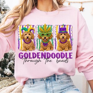 May include: Pink sweatshirt with a graphic of three golden doodles wearing Mardi Gras costumes. The dogs are in frames with purple, green, and yellow stripes. The text "GOLDENDOODLE Through The beads" is below the dogs.