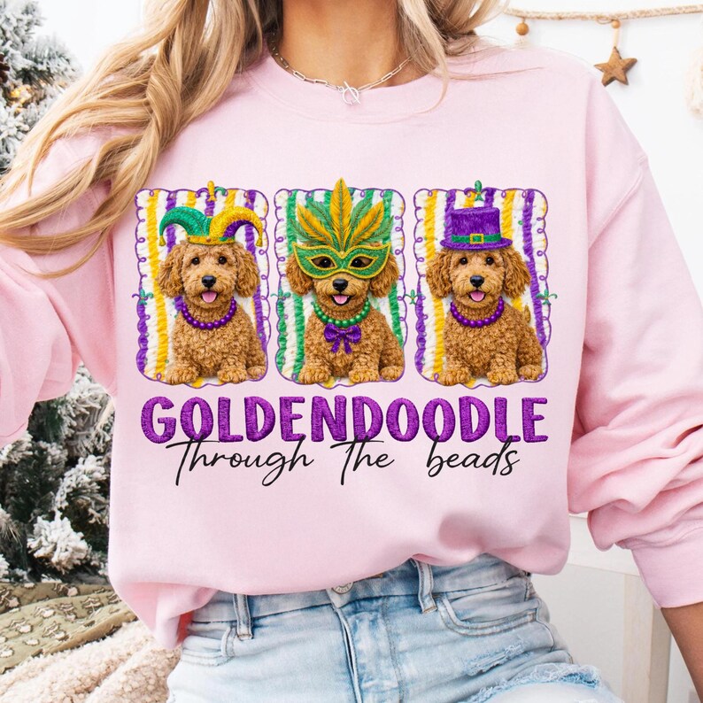 May include: Pink sweatshirt with a graphic of three golden doodles wearing Mardi Gras costumes. The dogs are in frames with purple, green, and yellow stripes. The text "GOLDENDOODLE Through The beads" is below the dogs.