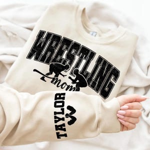 May include: A cream-colored sweatshirt with the words "WRESTLING Mom" in a black, distressed font. The design includes two wrestling figures. The sleeve displays the name "TAYLOR" in black, with a weightlifting icon.