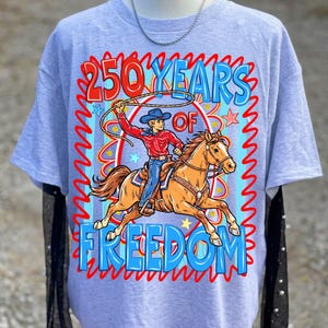 Puede incluir: Camiseta gris claro con un vaquero a caballo, con un lazo en la mano. El dise&ntilde;o incluye el texto "250 YEARS OF FREEDOM" en rojo y azul, con un borde decorativo. El vaquero lleva una camisa roja y vaqueros azules.