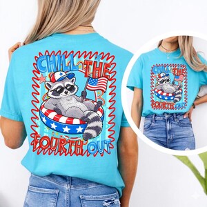 Op de afbeelding: Een lichtblauw t-shirt met een afbeelding van een wasbeer met een zonnebril en een patriottische hoed, ontspannen in een rood, wit en blauw opblaasbare ring. De tekst "Chill the Fourth Out" en een Amerikaanse vlag zijn zichtbaar.