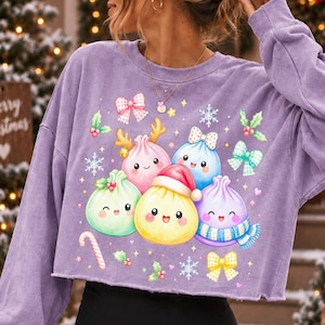 May include: A light purple long-sleeve sweatshirt featuring a graphic print of five smiling, colourful dumpling characters. The central yellow dumpling wears a red Santa hat, surrounded by snowflakes, holly berries, bows, and a candy cane.