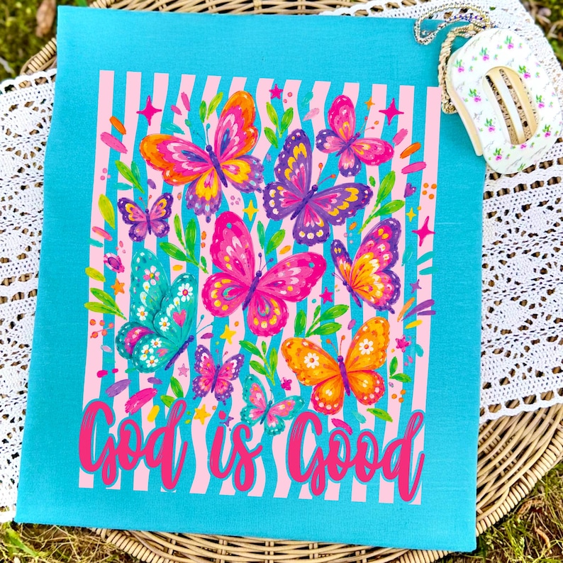 May include: A bright turquoise t-shirt featuring a central graphic of colorful butterflies in shades of pink, orange, purple, and teal over a light pink and white vertical striped background, with the text God is Good in a pink script font.