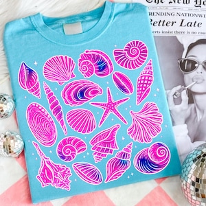 Pu&ograve; includere: Una maglietta azzurra con illustrazioni di conchiglie rosa e viola. Il design include varie forme di conchiglie e una stella marina, creando un'estetica a tema spiaggia. La maglietta ha un girocollo classico.