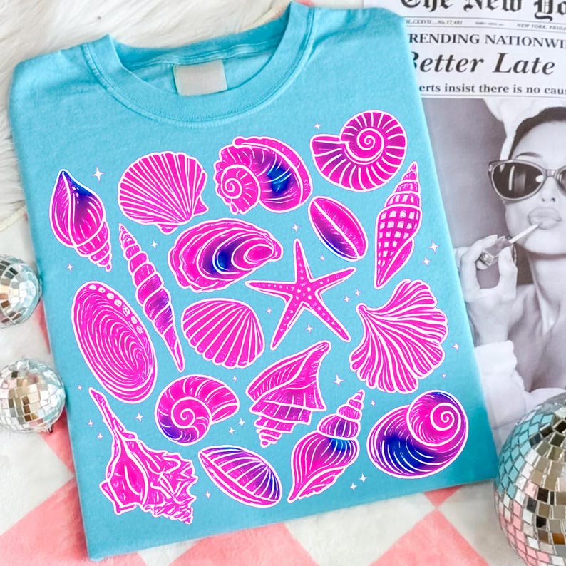 Pu&ograve; includere: Una maglietta azzurra con illustrazioni di conchiglie rosa e viola. Il design include varie forme di conchiglie e una stella marina, creando un'estetica a tema spiaggia. La maglietta ha un girocollo classico.