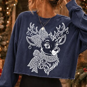 May include: A navy blue long-sleeve cropped sweatshirt featuring a white graphic print of a reindeer. The reindeer has a leopard print bow on its head and a matching leopard print scarf around its neck, with small ornaments hanging from its antlers.