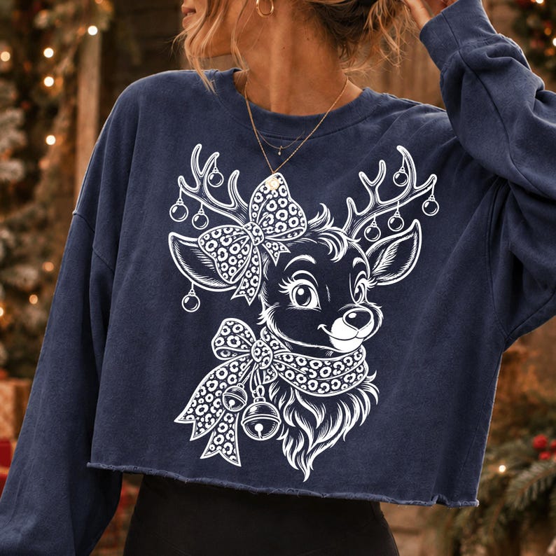 May include: A navy blue long-sleeve cropped sweatshirt featuring a white graphic print of a reindeer. The reindeer has a leopard print bow on its head and a matching leopard print scarf around its neck, with small ornaments hanging from its antlers.
