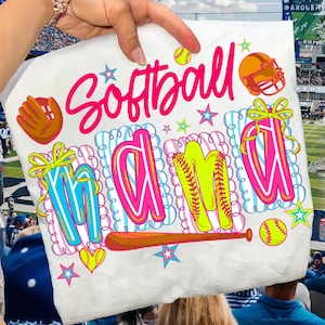 Könnte beinhalten: Weißes T-Shirt mit den Worten "Softball Mama" in Pink, Blau und Gelb. Das Design umfasst einen Softball, einen Handschuh, einen Helm, Sterne und einen Baseballschläger. Das T-Shirt wird von einer Person gehalten.