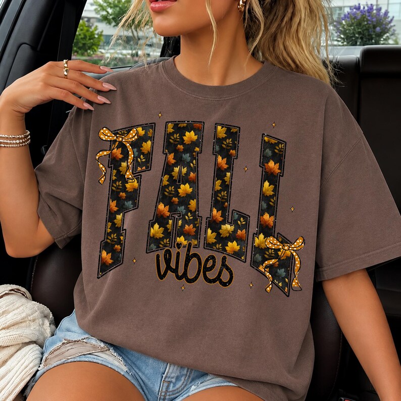 May include: A brown short-sleeved t-shirt featuring the text FALL in large block letters filled with a pattern of orange and yellow autumn leaves. The word vibes is written below in a cursive font. Two yellow polka-dot bows accent the design.