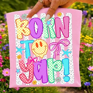 May include: Pink t-shirt featuring the phrase "BORN TO YAP!" in vibrant, patterned lettering. The design incorporates a smiley face, bows, stars, and flowers, set against a backdrop of white stripes and black dots.