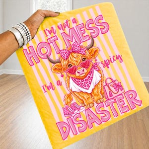 K&ouml;nnte beinhalten: Eine gelbe Decke mit rosa Streifen und dem Text "I'm not a HOT MESS, I'm a DISASTER." Eine Cartoon-Hochlandkuh mit herzf&ouml;rmiger Sonnenbrille, Schleife und Bandana ist abgebildet. Rosa Turnschuhe und eine Handtasche sind ebenfalls zu sehen.