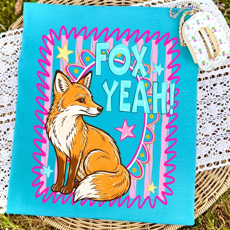 Op de afbeelding: Een turquoise vierkant textiel met een cartoon vos illustratie en de woorden "FOX YEAH!" in turquoise. De vos is oranje en wit, omringd door roze en gele sterren en een roze golvende rand.