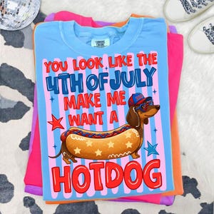 May include: A light blue t-shirt with the text "You look like the 4th of July make me want a hotdog." The graphic features a dachshund wearing sunglasses and a hat, designed as a hotdog. The shirt is stacked with other colorful shirts.
